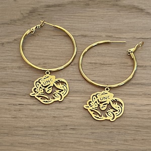 May include: Gold hoop earrings with a UNC Tar Heel logo charm. The logo is a stylized silhouette of a Tar Heel with the letters UNC inside.