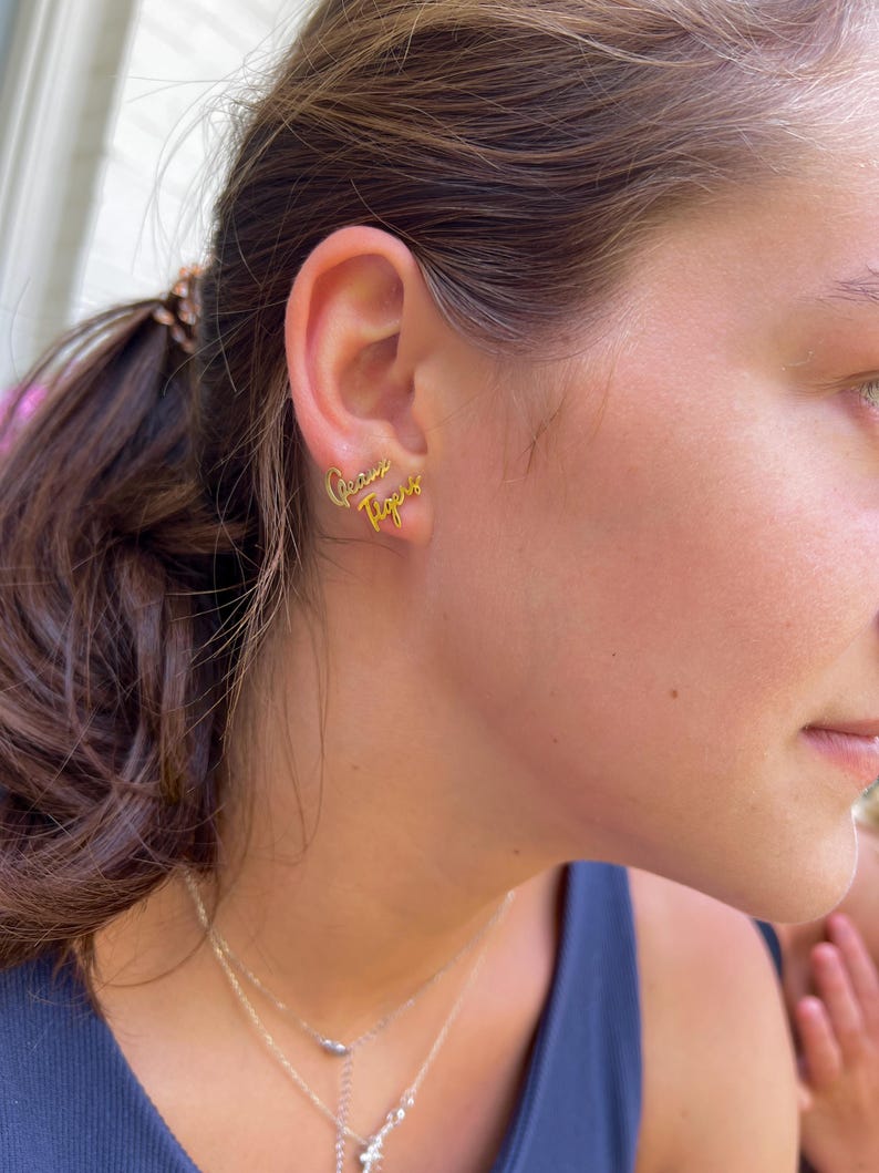 May include: Gold-colored stud earrings spelling out "Geaux Tigers" in a cursive font. The earrings are worn on an ear, with a person's face and neck visible in the background. Several necklaces are also visible.