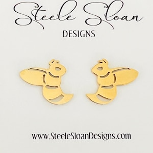 May include: A pair of gold-colored bee-shaped stud earrings. The earrings are cut-out designs with a stylized bee shape. The background is white with the text "Steele Sloan DESIGNS" and the website address www.SteeleSloanDesigns.com.