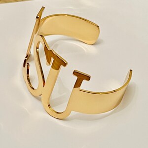 May include: A gold-colored cuff bracelet with the letters "LV" in a stylized font. The bracelet has a curved design and is made of a shiny metal. The bracelet is displayed on a white surface.
