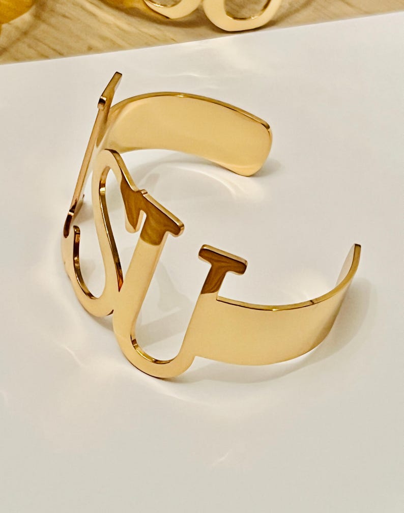 May include: A gold-colored cuff bracelet with the letters "LV" in a stylized font. The bracelet has a curved design and is made of a shiny metal. The bracelet is displayed on a white surface.