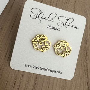 Może przedstawiać: Para złotych kolczyk&oacute;w z delikatnym wzorem kwiatowym i literami "UNC" w środku. Kolczyki są prezentowane na białej kartce z tekstem "Steele Sloan Designs" i adresem strony internetowej.
