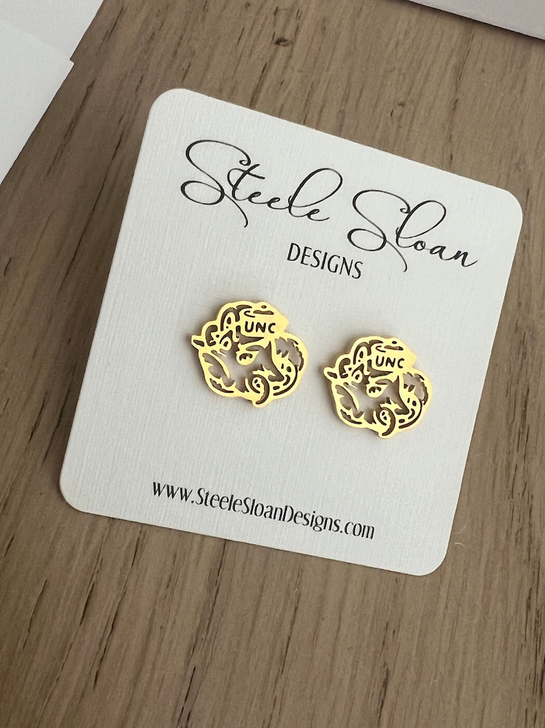 Może przedstawiać: Para złotych kolczyk&oacute;w z delikatnym wzorem kwiatowym i literami "UNC" w środku. Kolczyki są prezentowane na białej kartce z tekstem "Steele Sloan Designs" i adresem strony internetowej.