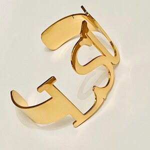 May include: A gold-colored cuff bracelet with an open design. The bracelet features the letters "LS" in a stylized font. The metal has a polished, reflective surface, and the bracelet is set against a white background.