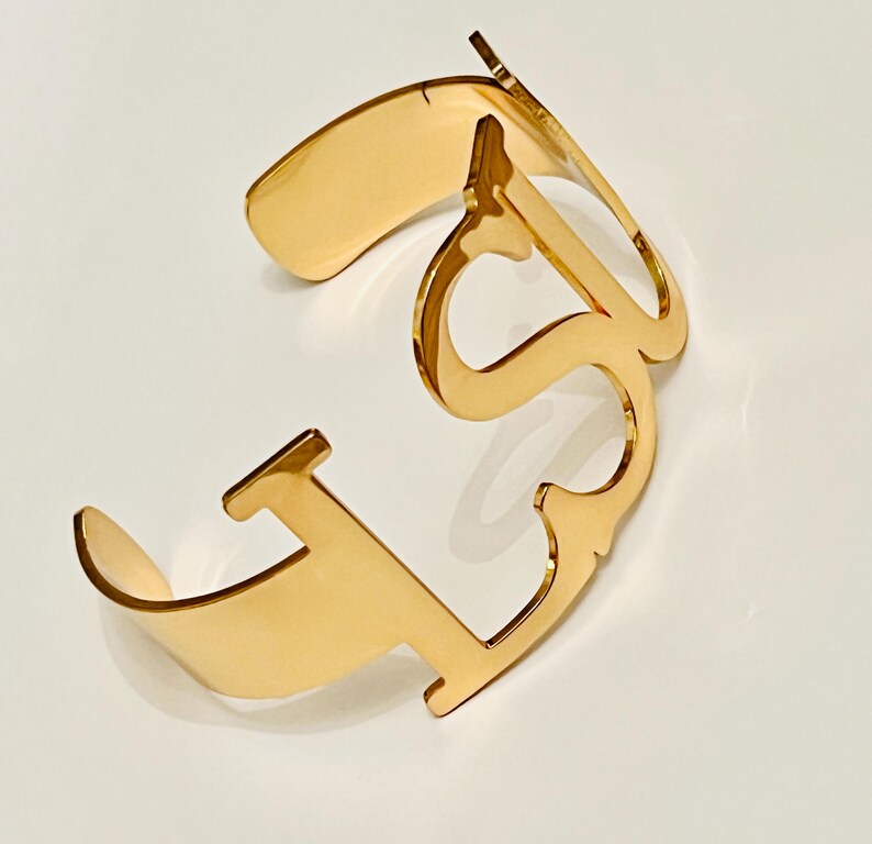 May include: A gold-colored cuff bracelet with an open design. The bracelet features the letters "LS" in a stylized font. The metal has a polished, reflective surface, and the bracelet is set against a white background.
