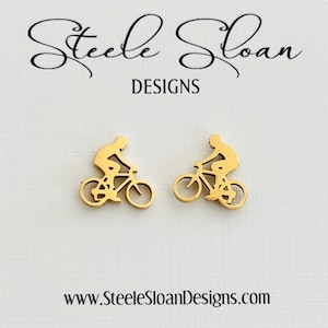 May include: A pair of gold-toned stud earrings featuring a silhouette of a person riding a bicycle. The earrings are on a white card with the text "Steele Sloan Designs" and "www.SteeleSloanDesigns.com".