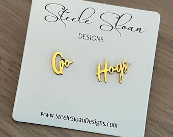 Può includere: Un paio di orecchini d'oro con le parole "Gro" e "Hogs" scritte in corsivo. Gli orecchini sono esposti su una carta bianca con il testo "Steele Sloan Designs" e un indirizzo di sito web.