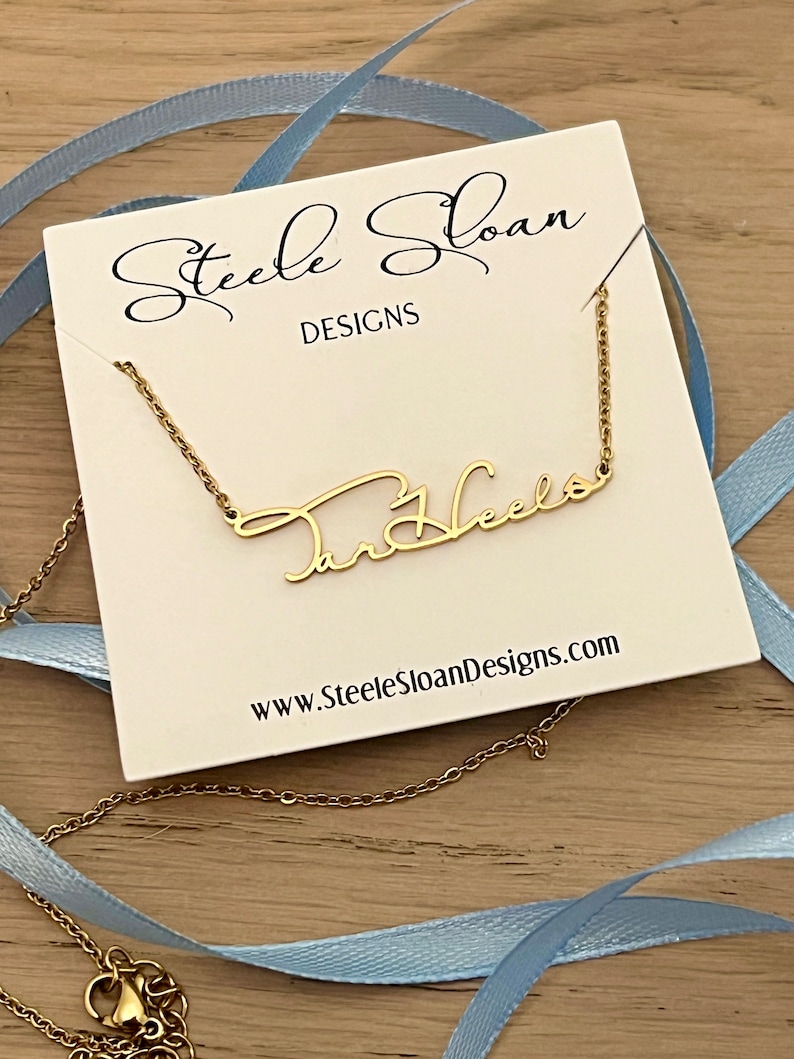 May include: A gold necklace with the name "Tar Heels" written in cursive script on a white card with the text "Steele Sloan Designs" and the website address "www.SteeleSloanDesigns.com".