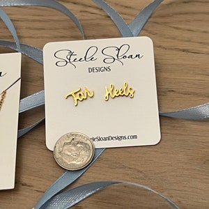 Może przedstawiać: Złote kolczyki z napisem "For Heels" na białej kartce z tekstem "Steele Sloan Designs" i stroną internetową "steelesloandesigns.com".