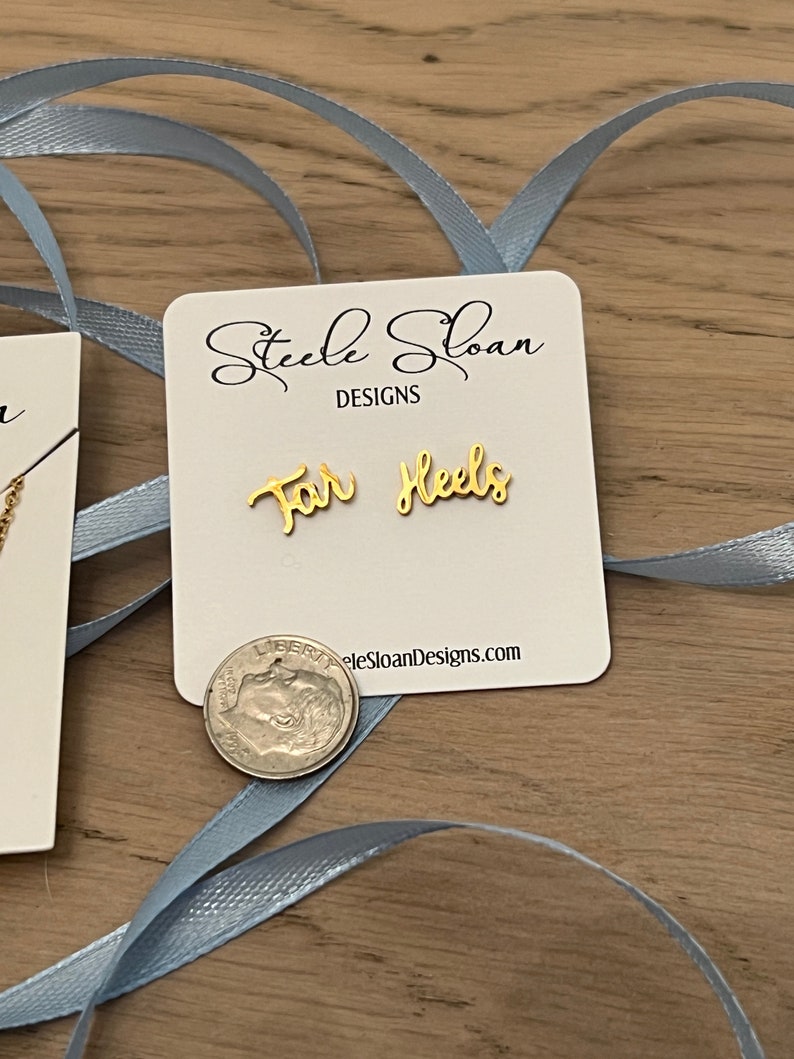 Może przedstawiać: Złote kolczyki z napisem "For Heels" na białej kartce z tekstem "Steele Sloan Designs" i stroną internetową "steelesloandesigns.com".