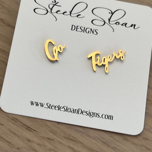 May include: Gold earrings with the words "Go Tigers" on them. The earrings are on a white card with the text "Steele Sloan Designs" and "www.SteeleSloanDesigns.com".