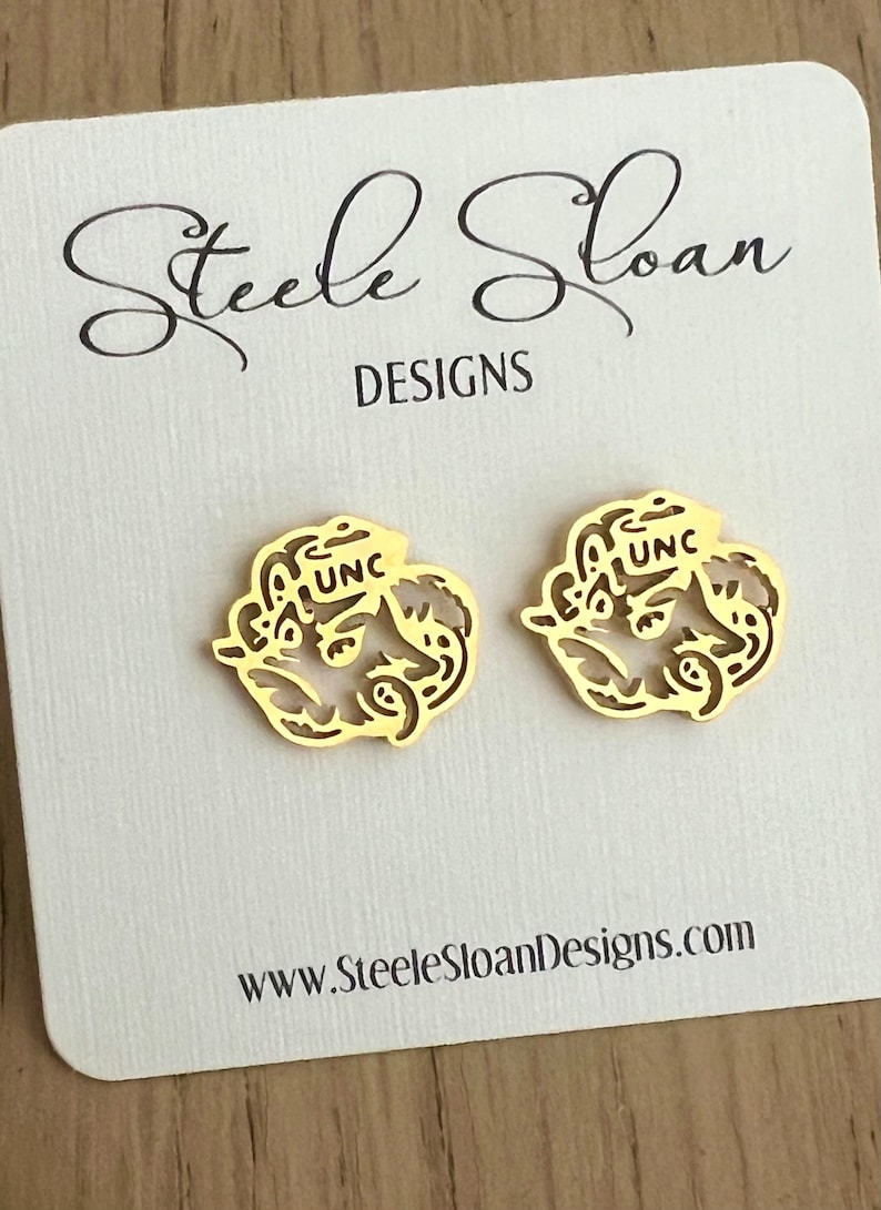 Może przedstawiać: Para złotych kolczyk&oacute;w z logo UNC. Kolczyki są prezentowane na białej kartce z tekstem "Steele Sloan Designs" i "www.SteeleSloanDesigns.com".