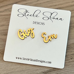 May include: Gold-colored earrings spelling out the names "Gigi" and "Em" on a white card. The card has the text "Steele Sloan Designs" and the website address www.SteeleSloanDesigns.com.