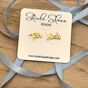 Może przedstawiać: Złote kolczyki z wygrawerowanym napisem "For Heels". Kolczyki znajdują się na białej kartce z tekstem "Steele Sloan Designs" i adresem strony internetowej.