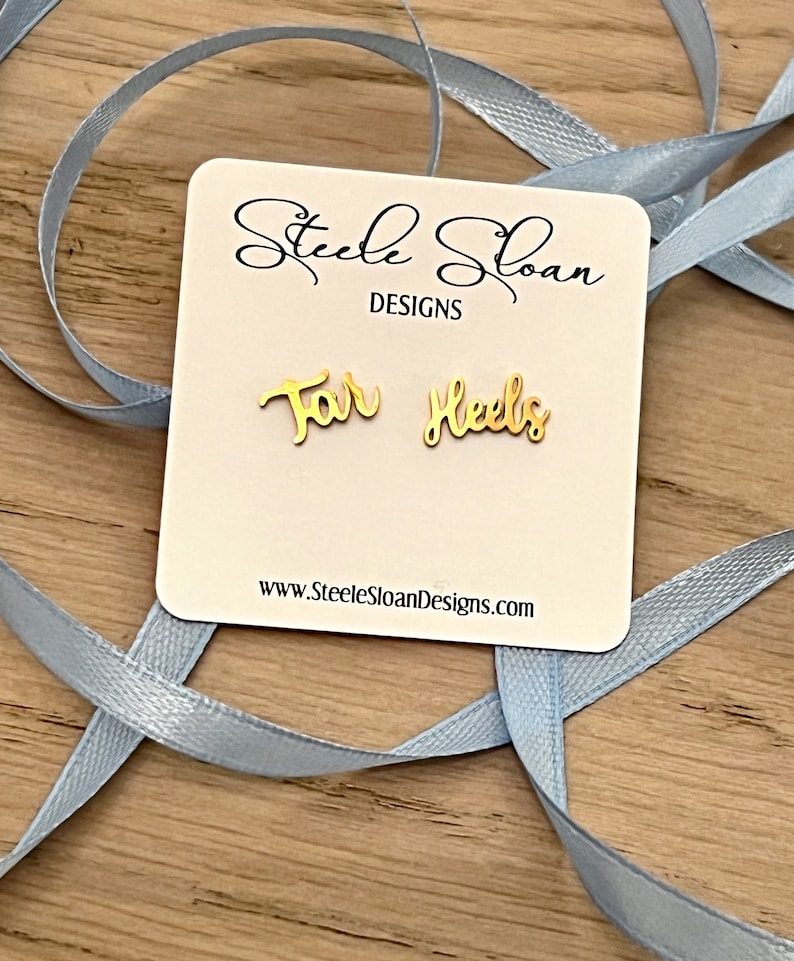 Może przedstawiać: Złote kolczyki z wygrawerowanym napisem "For Heels". Kolczyki znajdują się na białej kartce z tekstem "Steele Sloan Designs" i adresem strony internetowej.