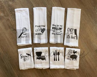 May include: Set of ten white kitchen towels with black text and graphics. The towels feature various kitchen-themed sayings, including "Just Beat It!" "It's Gettin' Hot In Here" "They See Me Rollin' They Hatin'" "Chop It Like It's Hot" "Screw It!" "Home" "Wakey Wakey Eggs & Bakey" and images of a whisk, a rolling pin, a chef's knife, a corkscrew, a house, and a rooster.