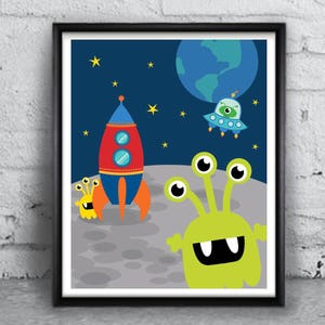May include: A framed print with a blue background featuring a rocket ship, a flying saucer, a green alien with three eyes, and a planet. The print is set against a white brick wall.