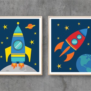 May include: Two framed prints with a space theme. The print on the left features a blue and yellow rocket with a porthole window. The print on the right features a red and blue rocket with a porthole window and a green planet in the background.