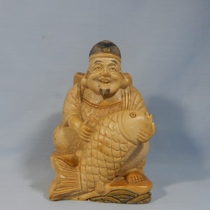 May include: A carved figurine of a smiling man holding a large fish. The figurine is made of a light brown material and has a detailed design.