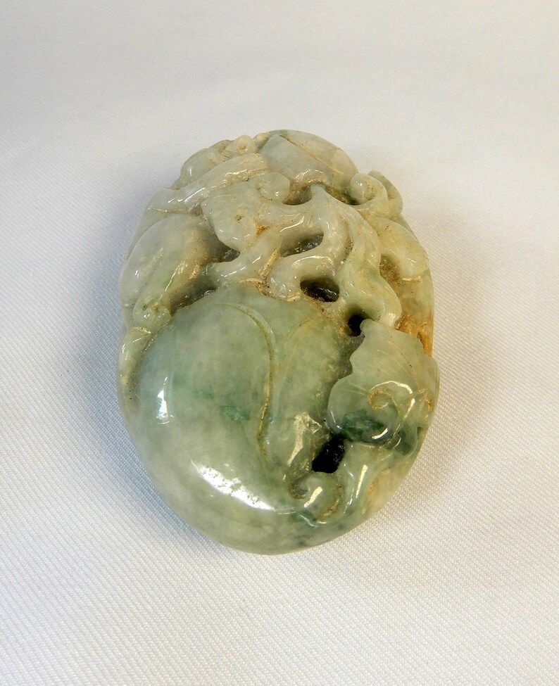 May include: A green jade carving of a mythical creature with intricate details. The carving is oval shaped and has a smooth, polished finish.