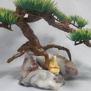 Op de afbeelding: Een miniatuur bonsai boomsculptuur met een bruine stam en groene bladeren. De boom staat op een veelkleurige rotsformatie, met een klein geel beeldje van een zittende figuur.
