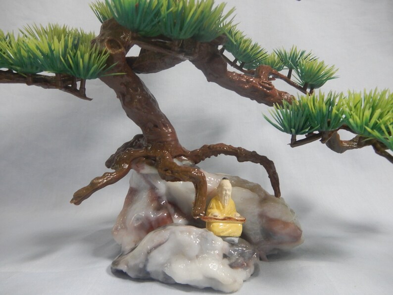 Op de afbeelding: Een miniatuur bonsai boomsculptuur met een bruine stam en groene bladeren. De boom staat op een veelkleurige rotsformatie, met een klein geel beeldje van een zittende figuur.
