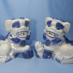 May include: Two ceramic guardian lion statues. The statues are white with blue accents, featuring detailed manes, expressive faces, and holding decorative spheres. The background is a solid blue.