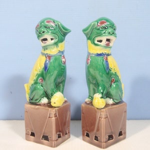 May include: A pair of ceramic Foo Dog figurines featuring a glossy green glaze with yellow accents on the mane and chest. Each creature stands on a brown square pedestal with cut-out details, one paw resting on a ball, with open mouths and ears.