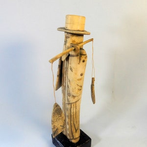 May include: A carved wooden figurine of a man wearing a top hat and carrying a fishing rod. The figure is painted in shades of beige and brown, with a fish and a small object hanging from the rod. The figurine is mounted on a black base.