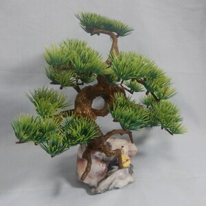 Op de afbeelding: Een miniatuur bonsai boom met heldergroene bladeren en een getextureerde bruine stam. De boom staat op een decoratieve rotsbasis met een klein beeldje. Deze kunstplant voegt een vleugje natuur toe aan elke ruimte.