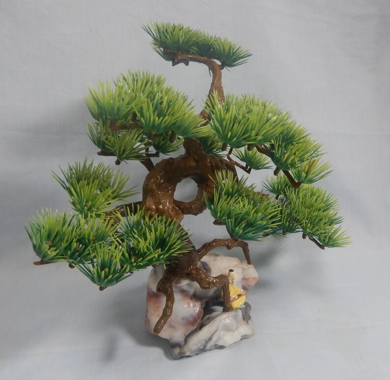 Op de afbeelding: Een miniatuur bonsai boom met heldergroene bladeren en een getextureerde bruine stam. De boom staat op een decoratieve rotsbasis met een klein beeldje. Deze kunstplant voegt een vleugje natuur toe aan elke ruimte.