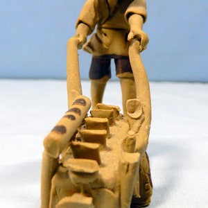 May include: A ceramic figurine of a person in a straw hat pushing a wheelbarrow with a brown handle and a clay base. The wheelbarrow is filled with small clay pots.