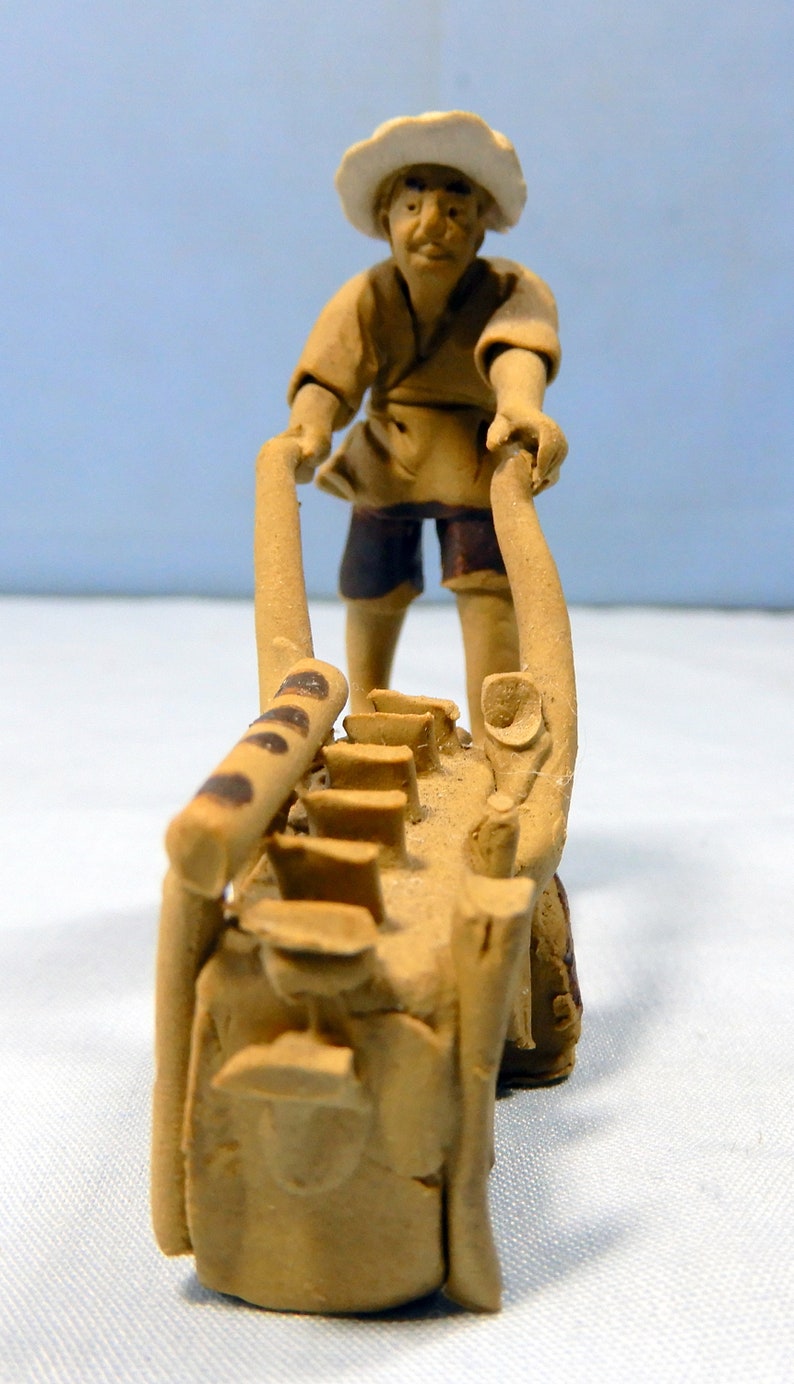 May include: A ceramic figurine of a person in a straw hat pushing a wheelbarrow with a brown handle and a clay base. The wheelbarrow is filled with small clay pots.