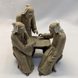 May include: A ceramic figurine depicting three elderly figures playing a board game. The figures are dressed in robes and have white hair and beards. The board game is on a small, round table. The figurine is a neutral tan color.