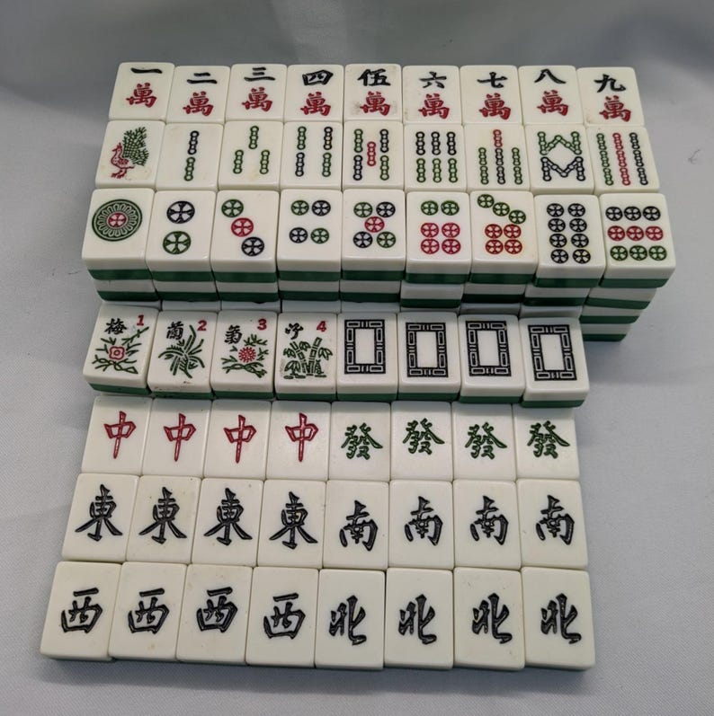 K&ouml;nnte beinhalten: Ein komplettes Set elfenbeinfarbener Mahjong-Steine mit gr&uuml;nen Kanten. Die Steine zeigen chinesische Schriftzeichen und Symbole in Rot und Schwarz. Das Set enth&auml;lt Steine mit Zahlen, Anz&uuml;gen und Sonderzeichen.