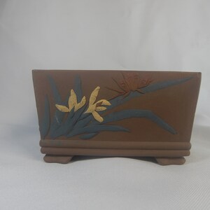May include: A rectangular brown ceramic planter with a floral design. The planter has a raised rim and four small feet. The design features yellow flowers, green leaves, and a red butterfly.