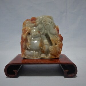 May include: A carved jade sculpture of a smiling Buddha figure with intricate details, in shades of orange, white, and green. The sculpture is mounted on a dark brown wooden stand, showcasing the artwork's form and color variations.