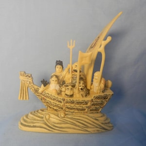 Peut inclure: Figurine ivoire représentant un bateau avec plusieurs personnages, un trident et une voile. Le bateau présente des sculptures détaillées et repose sur une base avec des motifs de vagues. Les personnages ont des traits de visage détaillés et sont vêtus de tenues traditionnelles.