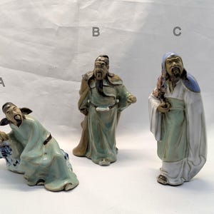 Op de afbeelding: Drie keramische beeldjes van Aziatische figuren in gewaden. De figuren zijn in tinten groen, wit en bruin. Eén figuur zit, een andere staat en de derde houdt een rol vast. De beeldjes hebben gedetailleerde gezichtskenmerken en lange baarden.