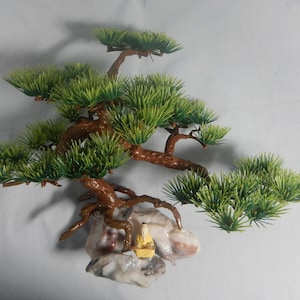Op de afbeelding: Een decoratieve bonsai-boom met groene bladeren en bruine takken. De boom is gemonteerd op een rotsformatie met een klein, geel beeldje. De achtergrond is een effen wit oppervlak.