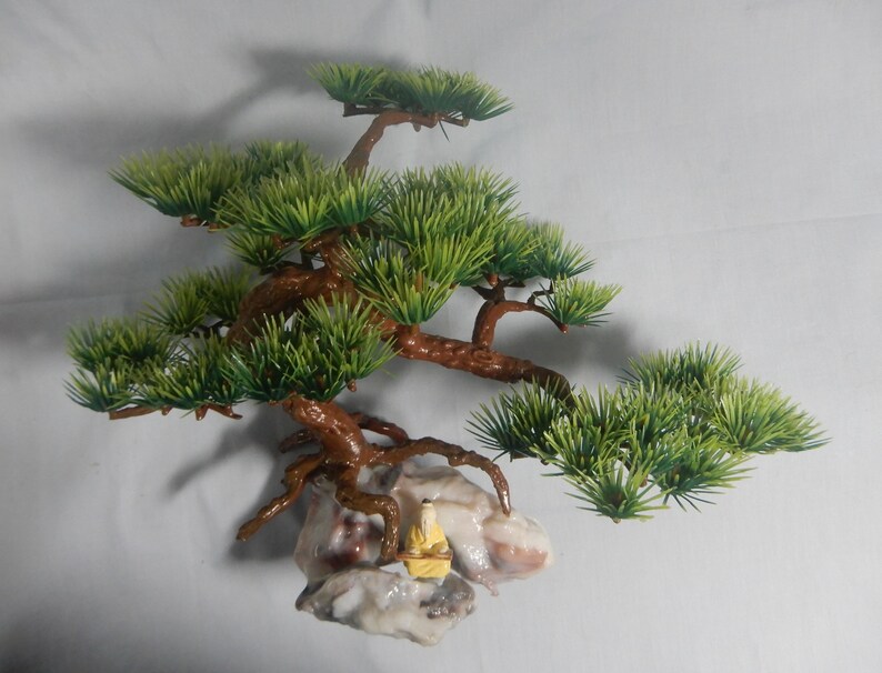 Op de afbeelding: Een decoratieve bonsai-boom met groene bladeren en bruine takken. De boom is gemonteerd op een rotsformatie met een klein, geel beeldje. De achtergrond is een effen wit oppervlak.