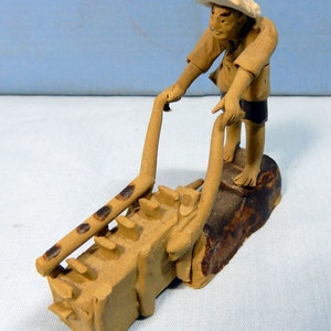 May include: A ceramic figurine of a person in a straw hat pushing a small, brown, clay cart with a ladder on the side.