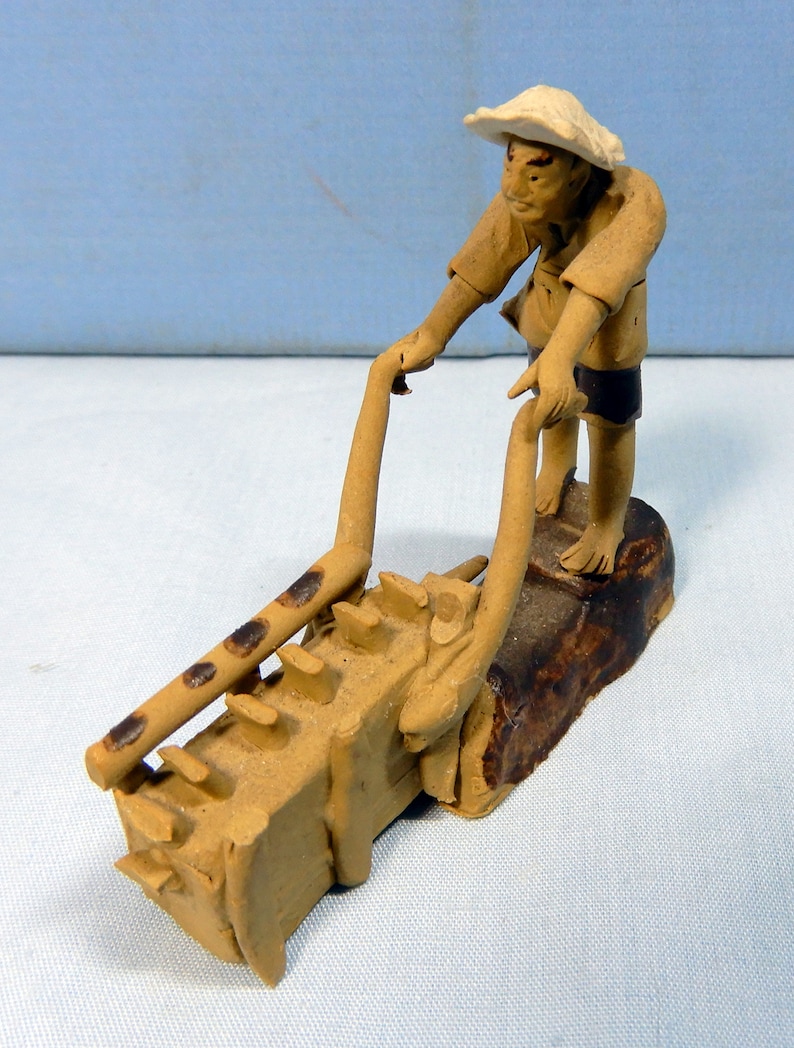May include: A ceramic figurine of a person in a straw hat pushing a small, brown, clay cart with a ladder on the side.
