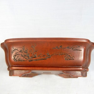 以下が含まれることがあります： 赤茶色の長方形の盆栽鉢。側面に風景画が彫られています。鉢は底が盛り上がっており、縁が湾曲しています。風景画には木、山、橋が描かれています。長さ約25cm。