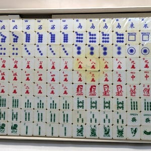 May include: A complete set of vintage Mahjong tiles, arranged in a box. The tiles are white with red, green, and blue markings, including Chinese characters and symbols. The set includes Jokers and other special tiles.