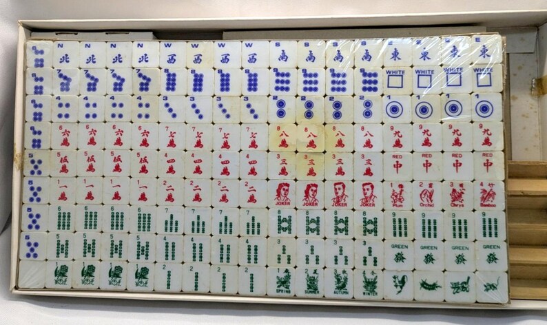 May include: A complete set of vintage Mahjong tiles, arranged in a box. The tiles are white with red, green, and blue markings, including Chinese characters and symbols. The set includes Jokers and other special tiles.