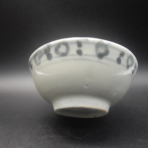 May include: A small, off-white ceramic bowl with a rounded shape. The rim is decorated with a repeating pattern of dark grey circles and dots. The bowl is set against a dark background.