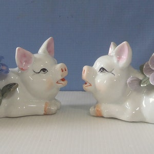 Könnte beinhalten: Zwei weiße Porzellan-Schweinefiguren mit lila und blauen Blumen. Die Schweine liegen mit hoch erhobenen Schnauzen.