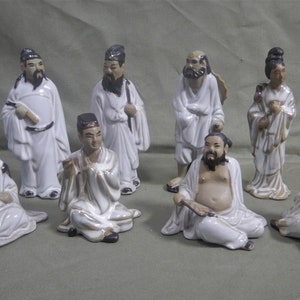 May include: Set of eight ceramic figurines depicting figures in traditional Chinese clothing. The figurines are painted in shades of white, brown, and black.