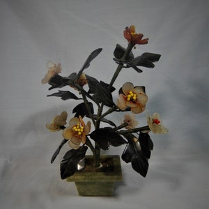 Puede incluir: Una planta de jade con hojas negras y flores hechas de ágata tallada. Las flores son una mezcla de blanco, marrón y naranja con centros amarillos. La planta está en una maceta de jade verde.